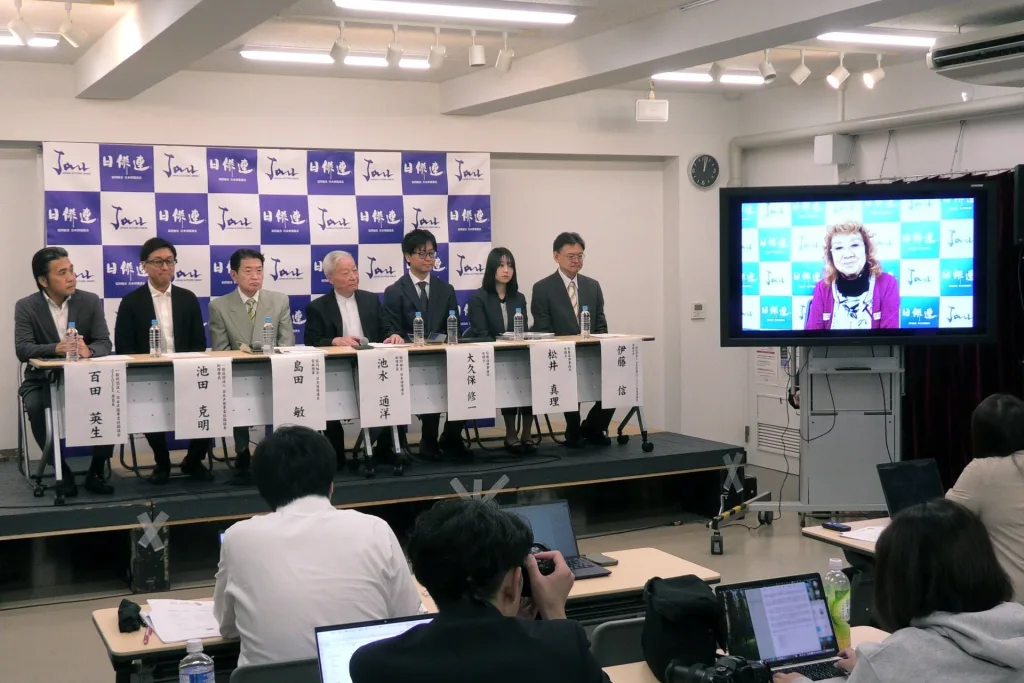 Seven speakers seated on a stage before a Japan Actors' Union backdrop at a press conference, with an actress on a video screen and reporters in the foreground