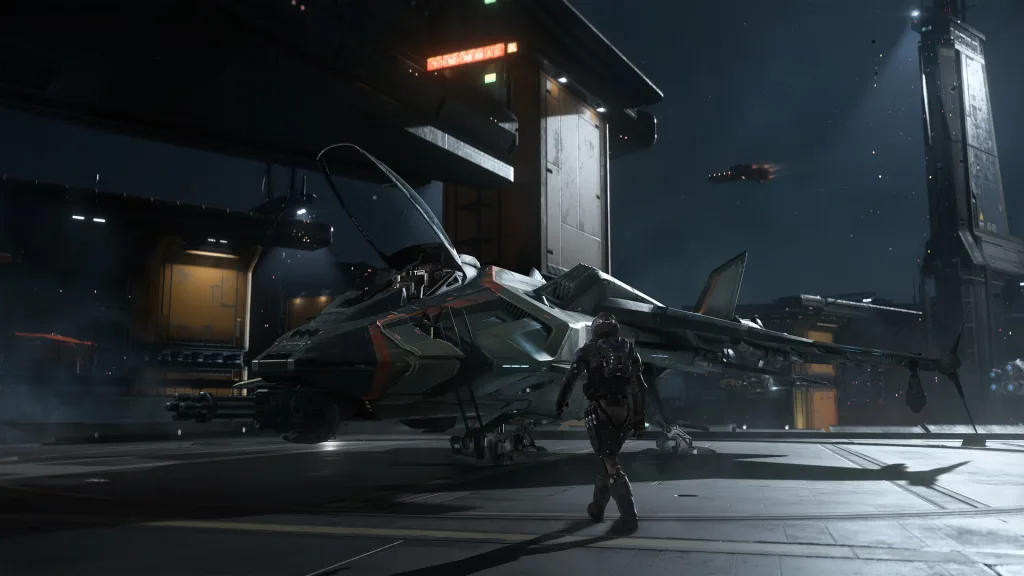 A pilot in a flight suit walks toward a dark grey fighter craft parked on a night-time hangar pad under floodlights.