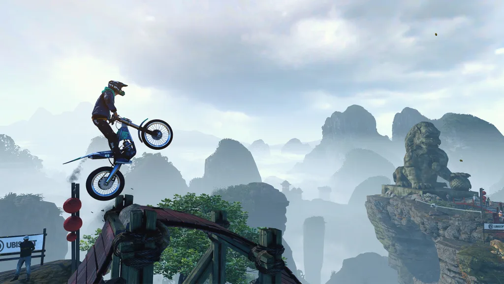 A trials rider on a blue motorbike leaps off a wooden ramp above misty karst mountains, with a stone lion statue on a cliff.
