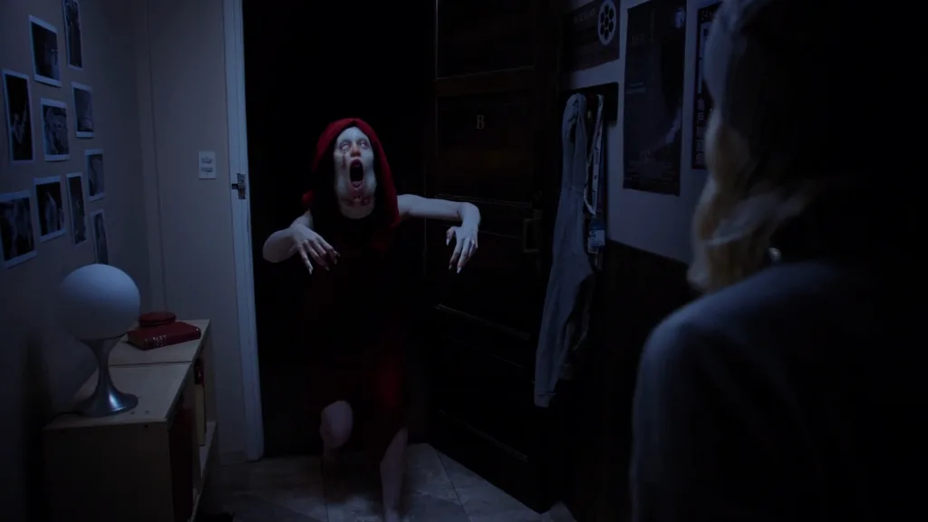 Live-action scene from Tenebris Somnia: a pale hooded creature lunges through a dark doorway at a blonde woman in the foreground