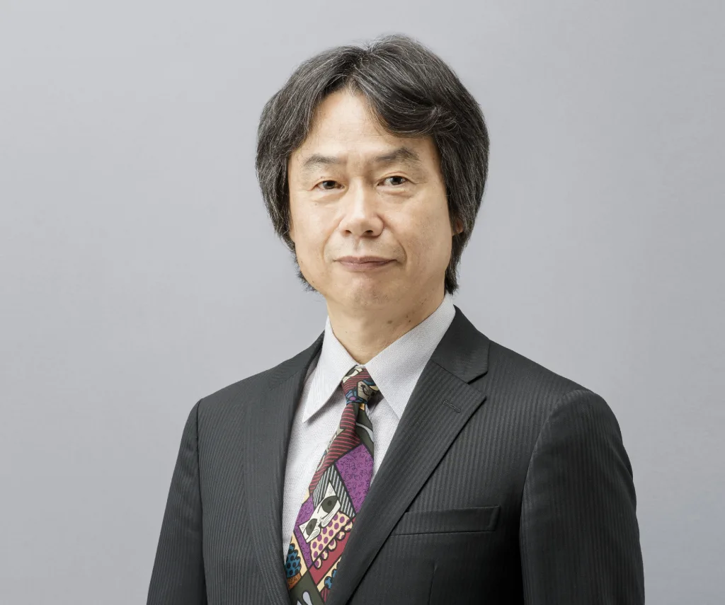Portrait of Shigeru Miyamoto in a dark suit and a patterned tie against a plain grey background.