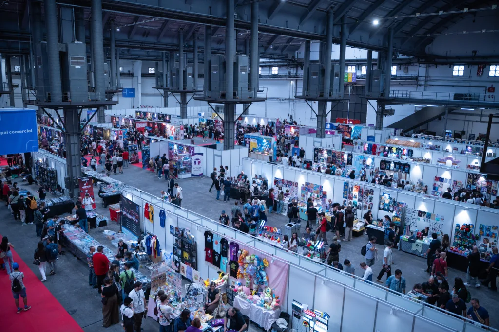 Aerial view of a busy exhibition hall with rows of white booths, merchandise stalls and crowds of visitors.